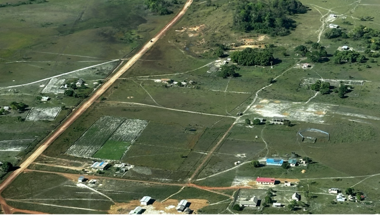 Aerial view of the red laterite Georgetown-Lethem Road running straight through Annai village in Guyana's North Rupununi, with houses, farm plots and forested hills beside it.